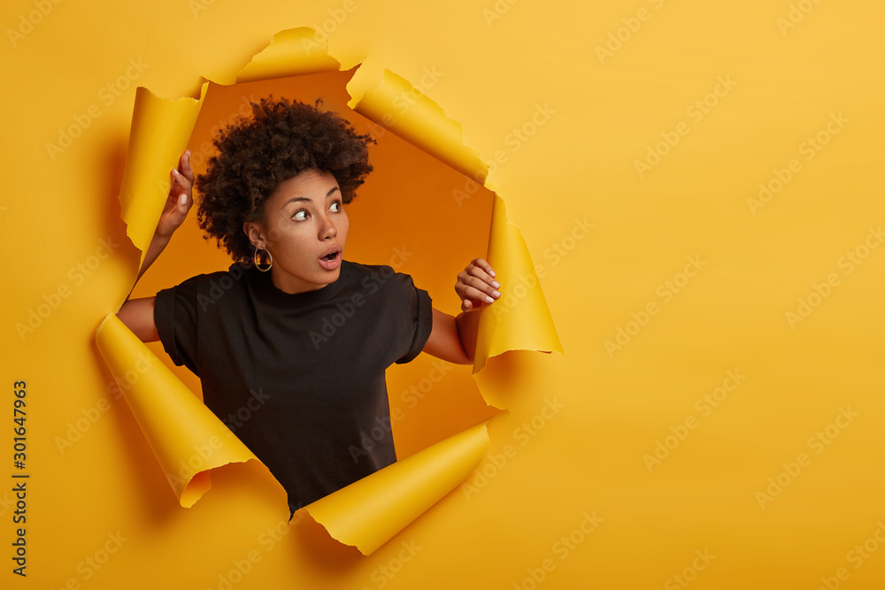 Photo of surprised Afro woman in black t shirt gasps from amazement, looks with scared face expression aside, dressed in black t shirt, unexpected to see something horrible, poses in torn paper wall - obrazy, fototapety, plakaty 