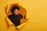 Fototapeta  - Photo of surprised Afro woman in black t shirt gasps from amazement, looks with scared face expression aside, dressed in black t shirt, unexpected to see something horrible, poses in torn paper wall