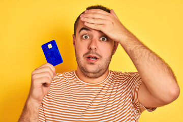 Sticker - Young man holding blue credit card standing over isolated yellow background stressed with hand on head, shocked with shame and surprise face, angry and frustrated. Fear and upset for mistake.