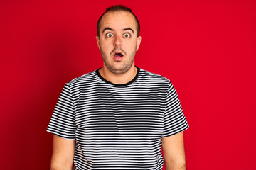 Sticker - Young man wearing striped navy t-shirt standing over isolated red background afraid and shocked with surprise expression, fear and excited face.