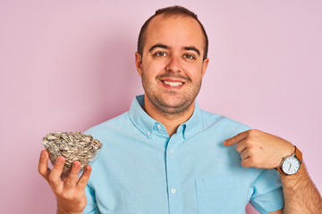 Sticker - Young man holding bowl with sunflowers seeds standing over isolated pink background with surprise face pointing finger to himself