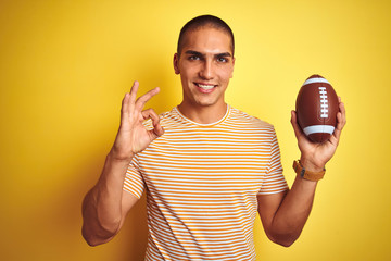 Poster - Young rugby player man holding a football ball over yellow isolated background doing ok sign with fingers, excellent symbol