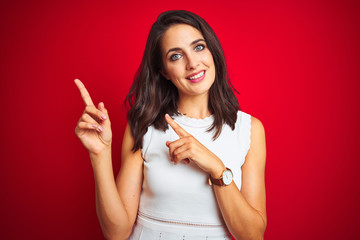 Young beautiful woman wearing white dress standing over red isolated background smiling and looking at the camera pointing with two hands and fingers to the side.