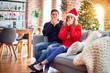 © Krakenimages.com - Young couple wearing santa claus hat sitting on the sofa around christmas tree at home shocked covering mouth with hands for mistake. Secret concept.