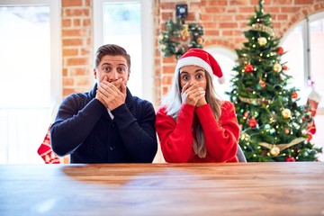 Canvas Print - Young couple wearing santa claus hat sitting on chair and table around christmas tree at home shocked covering mouth with hands for mistake. Secret concept.