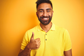 Young indian man wearing polo standing over isolated yellow background doing happy thumbs up gesture with hand. Approving expression looking at the camera with showing success.