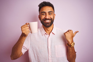 Poster - Young indian man wearing shirt drinking cup of coffee standing over isolated pink background pointing and showing with thumb up to the side with happy face smiling