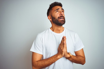 Canvas Print - Young indian man wearing t-shirt standing over isolated white background begging and praying with hands together with hope expression on face very emotional and worried. Asking for forgiveness