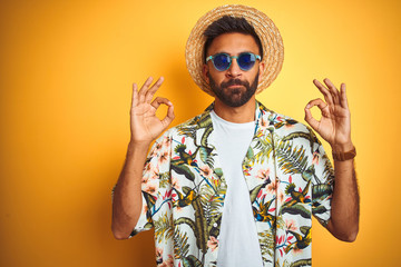 Wall Mural - Indian man on vacation wearing floral shirt hat sunglasses over isolated yellow background relax and smiling with eyes closed doing meditation gesture with fingers. Yoga concept.