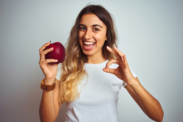 Poster - Young beautiful woman eating red apple over grey isolated background doing ok sign with fingers, excellent symbol