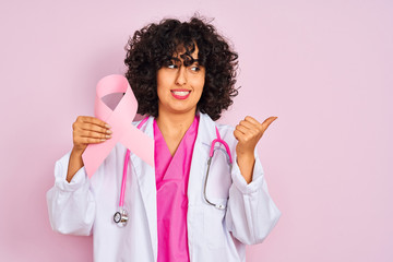 Sticker - Young arab doctor woman with curly hair holding cancer ribbon over isolated pink background pointing and showing with thumb up to the side with happy face smiling