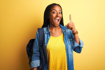 Wall Mural - African american student woman wearing backpack standing over isolated yellow background pointing finger up with successful idea. Exited and happy. Number one.