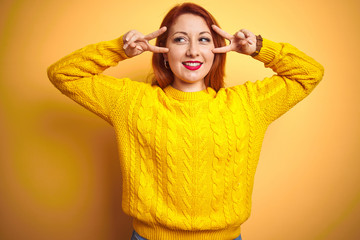 Sticker - Beautiful redhead woman wearing winter sweater standing over isolated yellow background Doing peace symbol with fingers over face, smiling cheerful showing victory