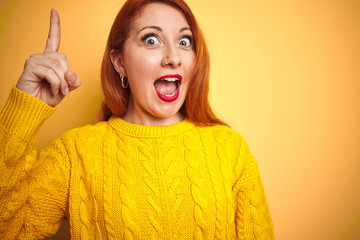 Wall Mural - Young redhead woman wearing winter sweater standing over isolated yellow background pointing finger up with successful idea. Exited and happy. Number one.