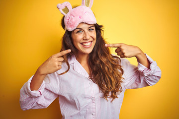 Wall Mural - Young woman wearing pajama and sleep mask standing over yellow isolated background smiling cheerful showing and pointing with fingers teeth and mouth. Dental health concept.