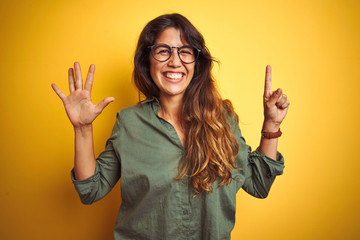 Wall Mural - Young beautiful woman wearing green shirt and glasses over yelllow isolated background showing and pointing up with fingers number six while smiling confident and happy.