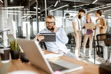 Senior confident manager working in the modern office with employees standing on the background. Concept of coworking and office work