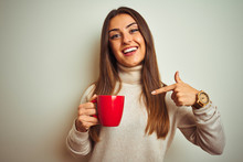 Young Beautiful Woman Drinking Red Cup Of Coffee Standing Over Isolated White Background With Surprise Face Pointing Finger To Himself