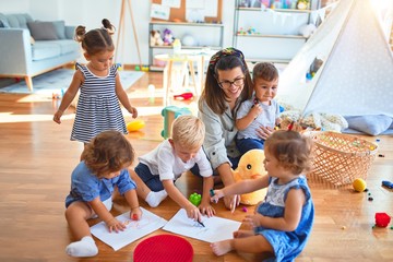 Wall Mural - Beautiful teacher and group of toddlers sitting on the floor drawing using paper and pencil around lots of toys at kindergarten