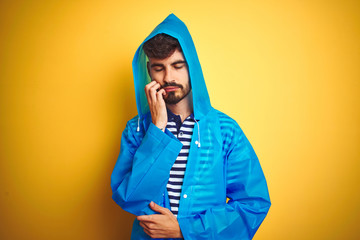 Wall Mural - Young handsome man wearing rain coat with hood standing over isolated yellow background thinking looking tired and bored with depression problems with crossed arms.
