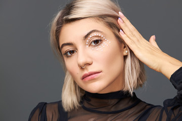 Beauty, style and femininity concept. Studio shot of gorgeous sensual European female with blonde bob hairstyle demonstrating her stylish make up, white crystals around eye, looking at camera