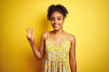 Sticker - African american woman wearing casual floral dress standing over isolated yellow background showing and pointing up with fingers number three while smiling confident and happy.