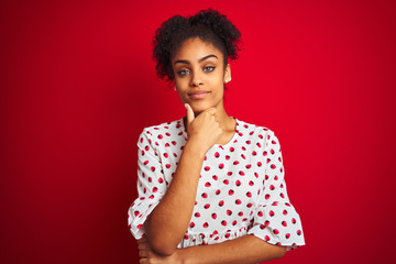 Sticker - African american woman wearing fashion white dress standing over isolated red background looking confident at the camera smiling with crossed arms and hand raised on chin. Thinking positive.