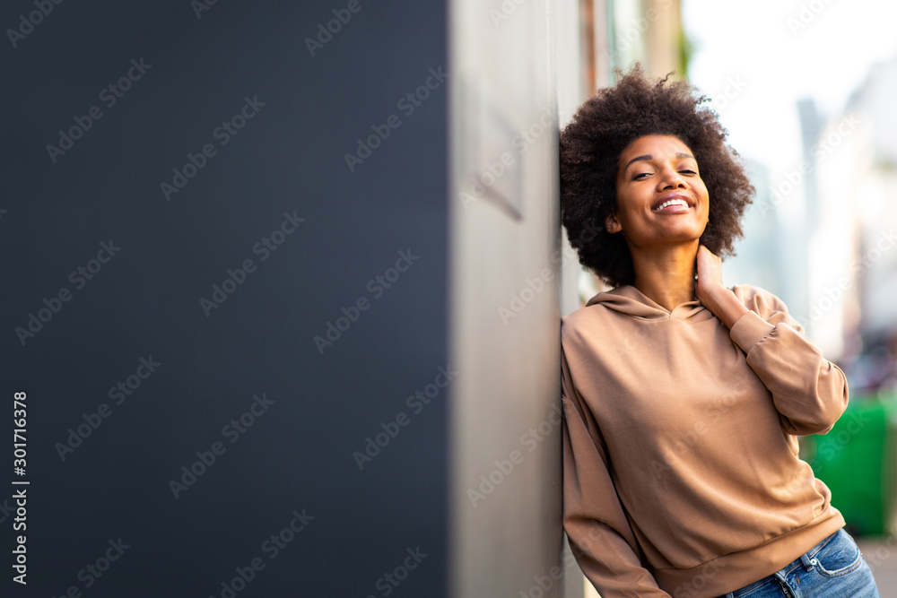 beautiful black female fashion model with afro hairstyle leaning against wall and smiling - obrazy, fototapety, plakaty 