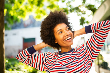 Wall Mural - Close up happy young black woman smiling with hands behind head in park and looking up