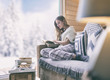 Young beautiful blonde woman with book sitting home in living room by the window. Winter snow landscape view.