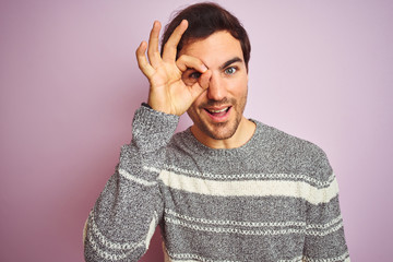 Canvas Print - Young handsome man wearing casual striped sweater standing over isolated pink background with happy face smiling doing ok sign with hand on eye looking through fingers