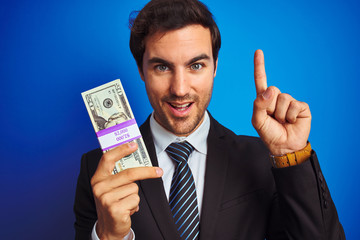 Poster - Young handsome businessman wearing suit holding dollars over isolated blue background surprised with an idea or question pointing finger with happy face, number one