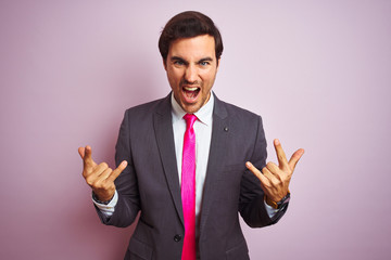 Wall Mural - Young handsome businessman wearing suit and tie standing over isolated pink background shouting with crazy expression doing rock symbol with hands up. Music star. Heavy concept.