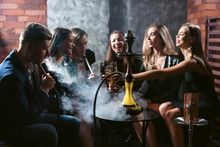 Friends Party In Hookah Lounge. Group Of People Women And Men Smoking Shisha In Cafe Or Bar, Making Smoke Clouds, Having Fun, Smiling. Relax Concept. Friendship