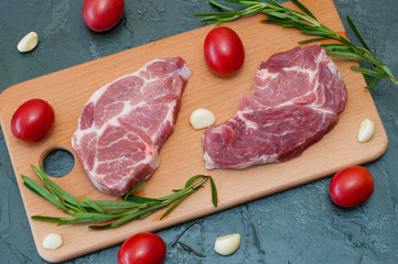 Flat lay, raw meat two pork steak with tomatoes garlic and rosemary on a wooden cutting board close-up