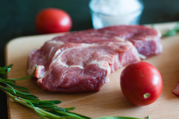 Wall Mural - Raw pork steak, salt, rosemary and tomato on cutting board, on a dark gray concrete background, close up, in dark tonality