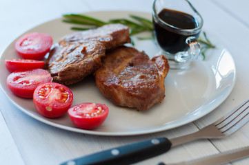 Wall Mural - Tasty, grilled pork steaks on a gray plate with sauce, rosemary and tomatoes, on light wooden background, cutlery, soft focus