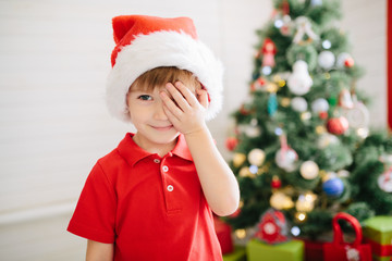 Wall Mural - Portrait of a little cute boy with blue eyes in a Santa Claus hat in a decorated Christmas room with a xmas tree.