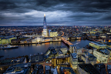 Fototapete - Blick auf die markante Stadtlandschaft von London am Abend mit bewölktem Himmel und beleuchteten Gebäuden entlang der Themse