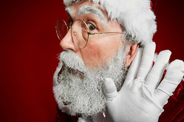 Wall Mural - Close up portrait of classic Santa Claus putting hand to ear trying to hear secrets while standing against red background