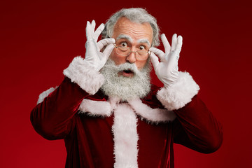 Wall Mural - Waist up portrait of surprised Santa Claus looking at camera and adjusting glasses while posing against red background in studio, copy space