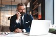 © opolja - Portrait of a nice smiling man with beard, he is sitting at table, talking on the phone with his laptop