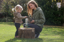 Mother And Her Toddler Girl Putting Seeds For Birds In A Bird Feeder. Quality Outdoor Family Time Together. Encouraging Wildlife In The Garden With Bird Feeder.
