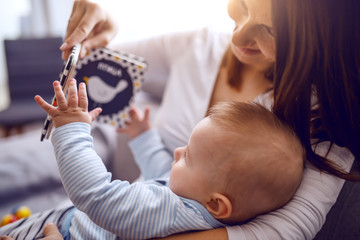 happy proud caucasian mother sitting on sofa in living room, holding her adorable son and showing hi