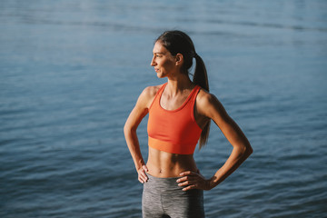 Wall Mural - Attractive sporty fit caucasian brunette in sportswear and with ponytail standing near water with hands on hips while looking away.