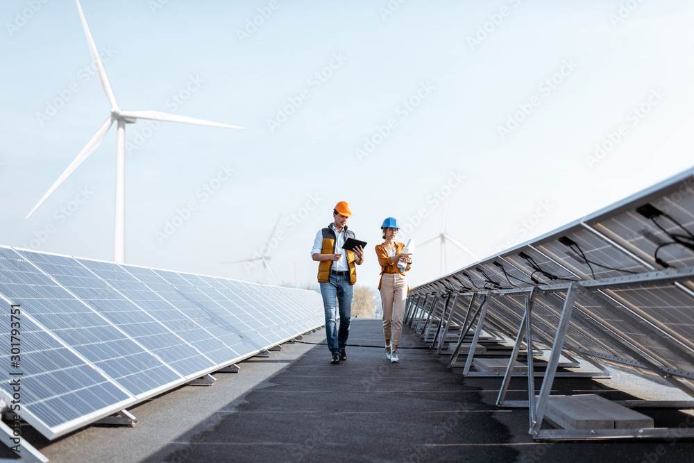 View on the rooftop solar power plant with two engineers walking and examining photovoltaic panels. Concept of alternative energy and its service - obrazy, fototapety, plakaty 
