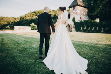 Wall Mural - Gorgeous bride and groom walking in sunset light. Beautiful stylish wedding couple embracing in summer park, back view. Sensual romantic moment. Newlyweds