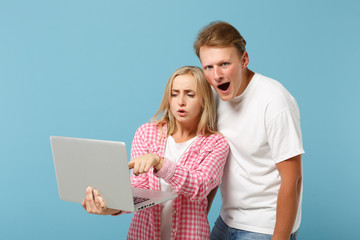 Young shocked couple friends guy girl in white pink empty blank t-shirts posing isolated on pastel blue background. People lifestyle concept. Mock up copy space. Pointing finger on laptop pc computer.