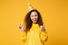 Cheerful Young African American Girl In Fur Sweater, Birthday Hat Posing Isolated On Yellow Orange Background. People Lifestyle Concept. Mock Up Copy Space. Holding Pipe, Showing Thumb Up, Blinking.