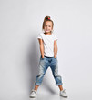 © Dmitry Lobanov - Happy smiling kid girl in white t-shirt and sneakers is posing with her hands in pockets of her torn jeans on white